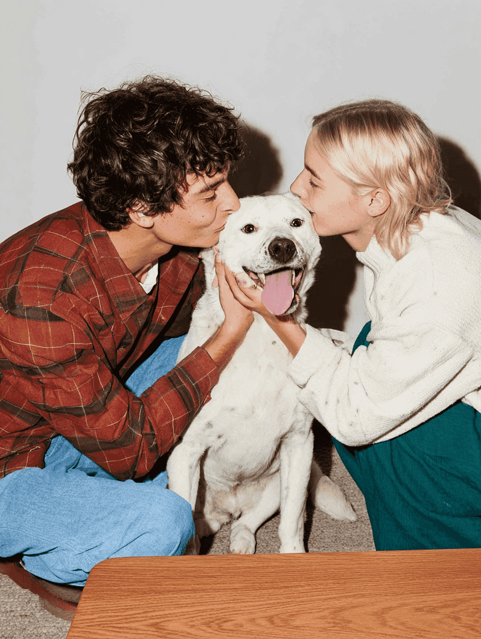 A young couple sit on the floor, each kissing a happy white dog on either cheek. The dog's tongue is out.